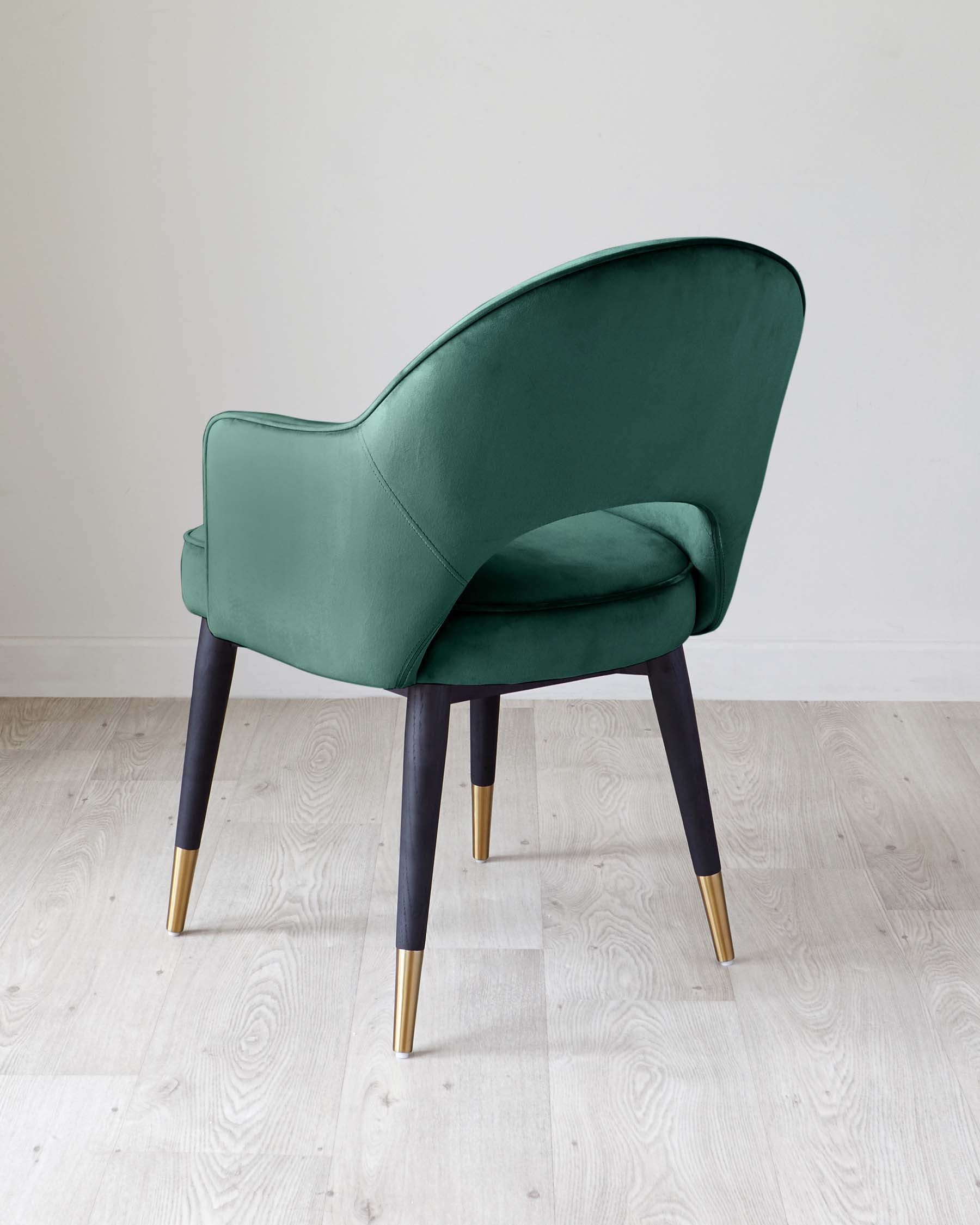 Elegant emerald green upholstered armchair with curved backrest and seat, featuring dark stained wooden legs with stylish gold-toned metal tips, positioned on a light hardwood floor against a neutral wall.