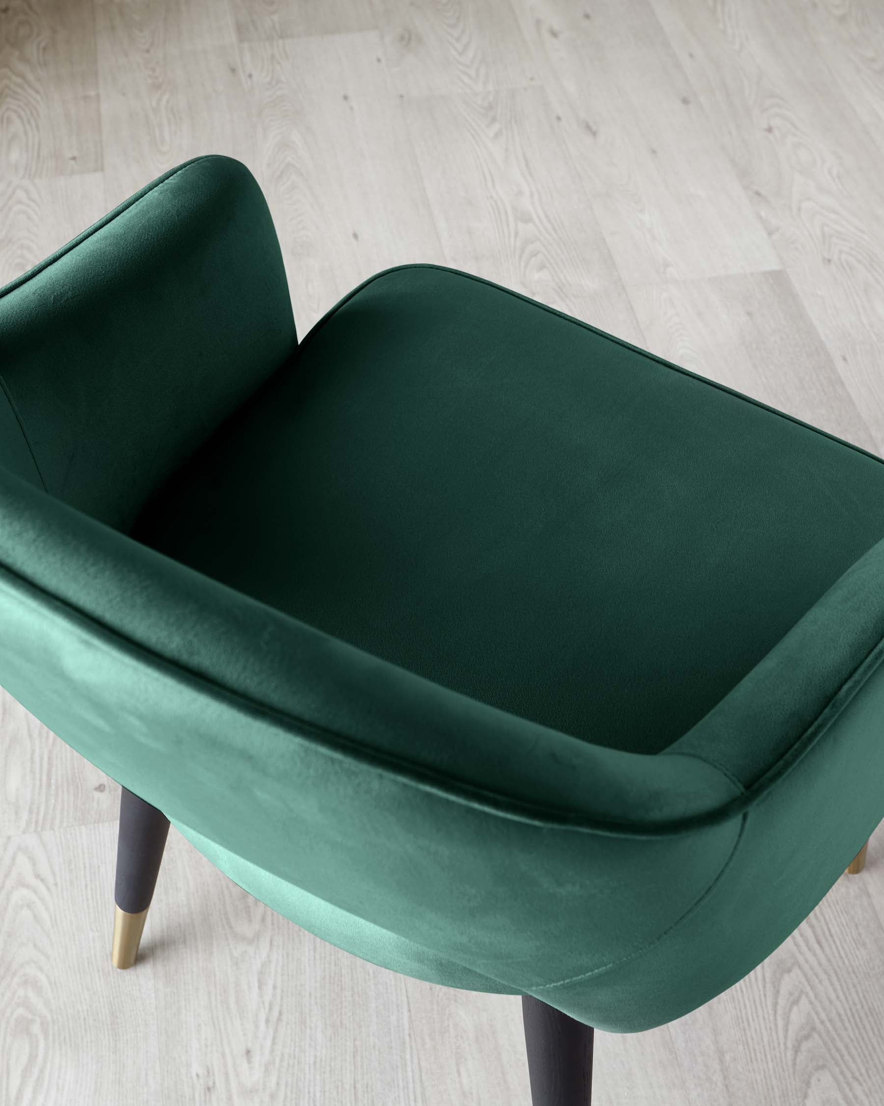 Contemporary, emerald green upholstered armchair with a smooth, curved backrest and comfortable, wide seat cushion. The chair features four tapered legs, the front two capped with brass tips, set against a light wooden floor.