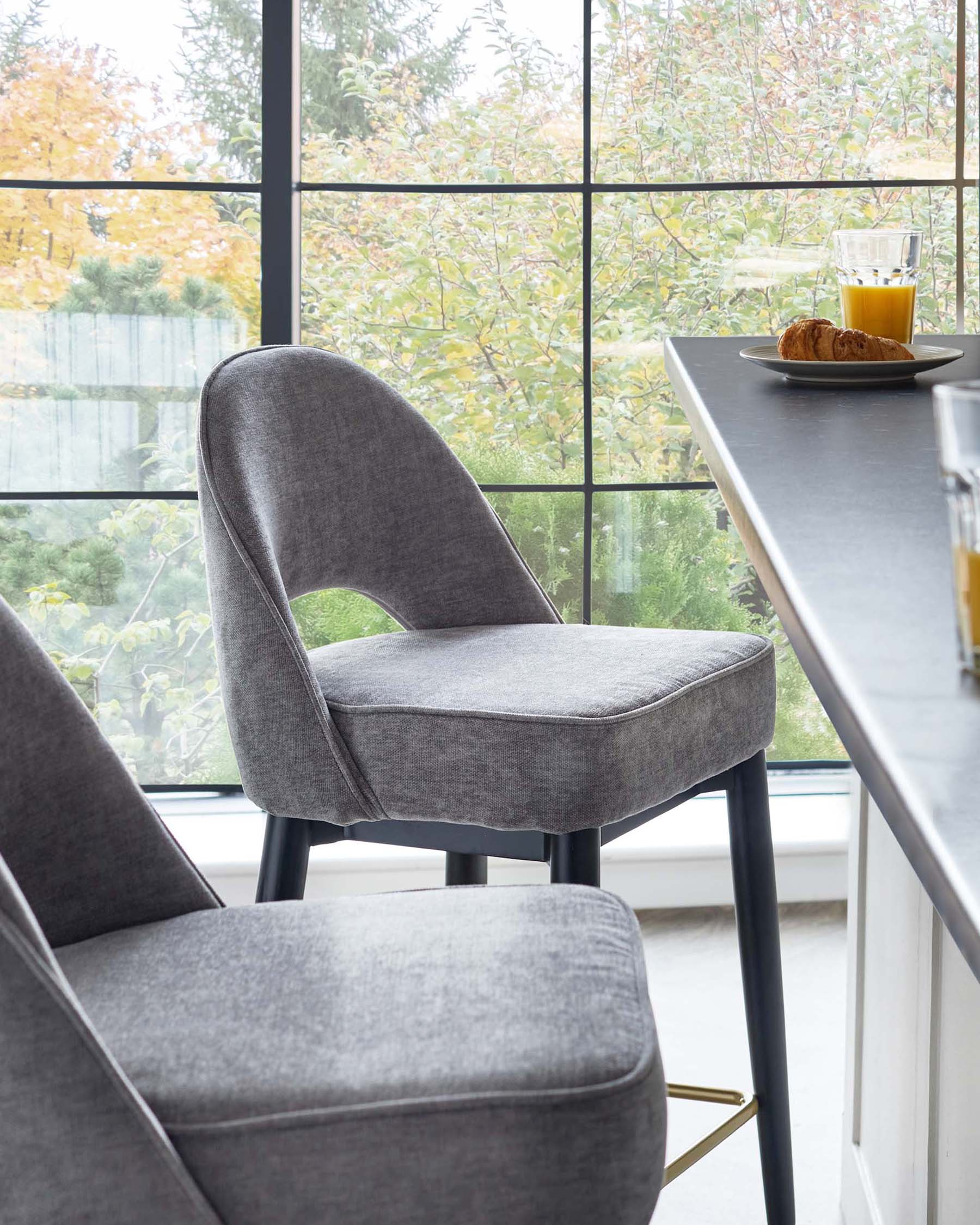 Modern upholstered dining chair in a dark grey fabric, featuring a curved backrest and a comfortable seat, with sleek black metal legs that include gold accents at the feet. A portion of a bar table is visible with a plate holding a croissant and a glass of orange juice on top, placed by a large window overlooking a garden.