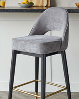 Elegant modern bar stool with a curved backrest, upholstered in grey fabric, featuring a padded seat for comfort. The chair has four black wooden legs which are connected by a square, gold-finished metal footrest, providing stability and a chic contrast.