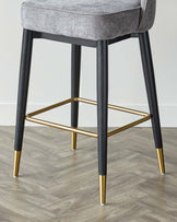 Modern bar stool with grey fabric cushioned seat and black tapered legs featuring gold-finished accents and footrest.