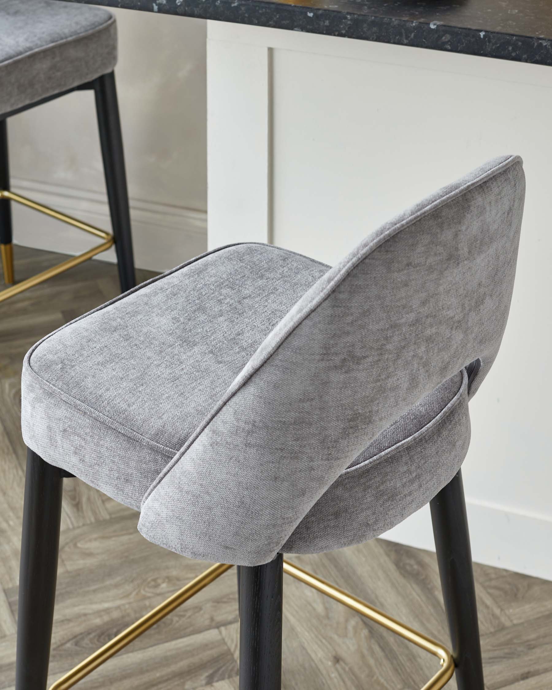 Elegant modern bar stool with a sleek black frame, featuring a curved design and gold footrest accents. The chair has a comfortable padded seat and backrest upholstered in a textured light grey fabric that creates a sophisticated contrast with the dark legs.