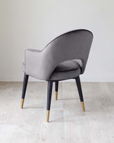 Elegant contemporary chair with a smooth grey velvet upholstery and a curved backrest, complemented by dark wooden legs tipped with brass accents. The chair is positioned on a light wooden floor against a neutral background.