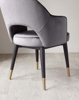 Elegant modern dining chair with grey velvet upholstery and dark wooden legs accented with gold tips, set against a minimalist background with light wooden flooring.