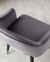 Modern charcoal grey accent chair with a smooth fabric finish, featuring a rounded backrest and a spacious seat. It has an understated design with four slanted legs in black, complemented by gold-tone tips, set on a light herringbone-pattern wood floor.