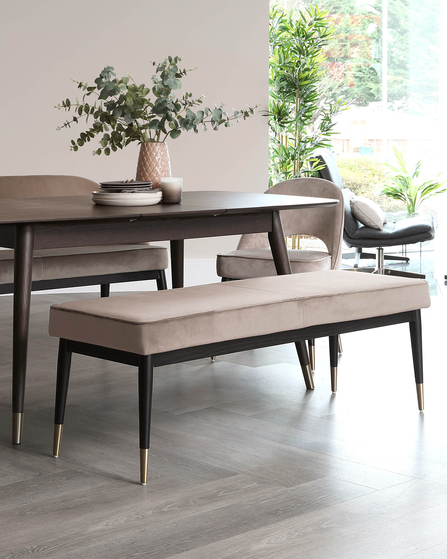 Contemporary dining room furniture including a dark wood rectangular dining table, accessorized with a vase of eucalyptus branches and simple tableware, complemented by taupe upholstered dining benches with black legs tipped in gold, and a matching armchair in the background, all set upon a hardwood floor.