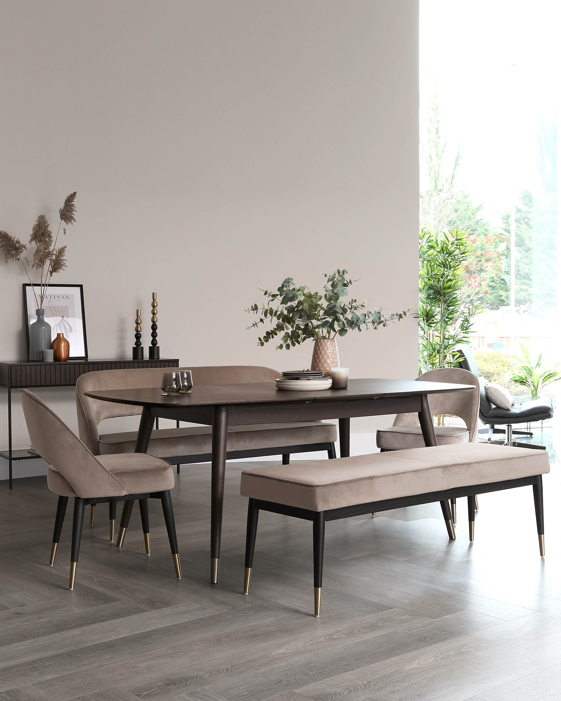 Elegant modern dining room set featuring a dark wood oval table with sleek tapered legs tipped with brass accents. Includes two plush, upholstered armchairs with matching wooden legs and brass details, and a coordinating bench with similar design attributes. The set conveys a sense of contemporary sophistication suitable for a stylish dining space.