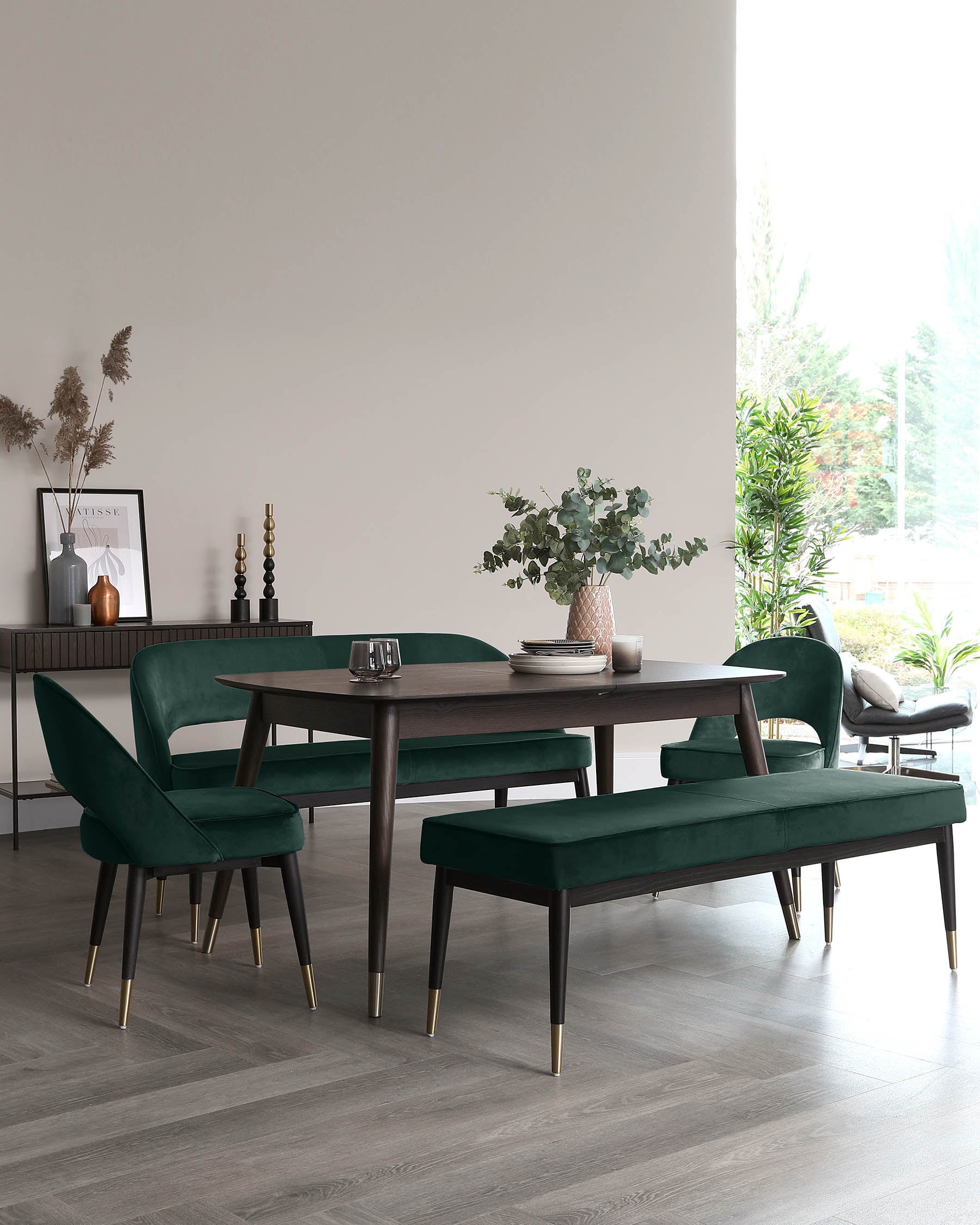 Modern dining set featuring a dark wood table with gold-tipped legs, complemented by four plush emerald green velvet chairs and a matching bench, also with gold accents on the legs.