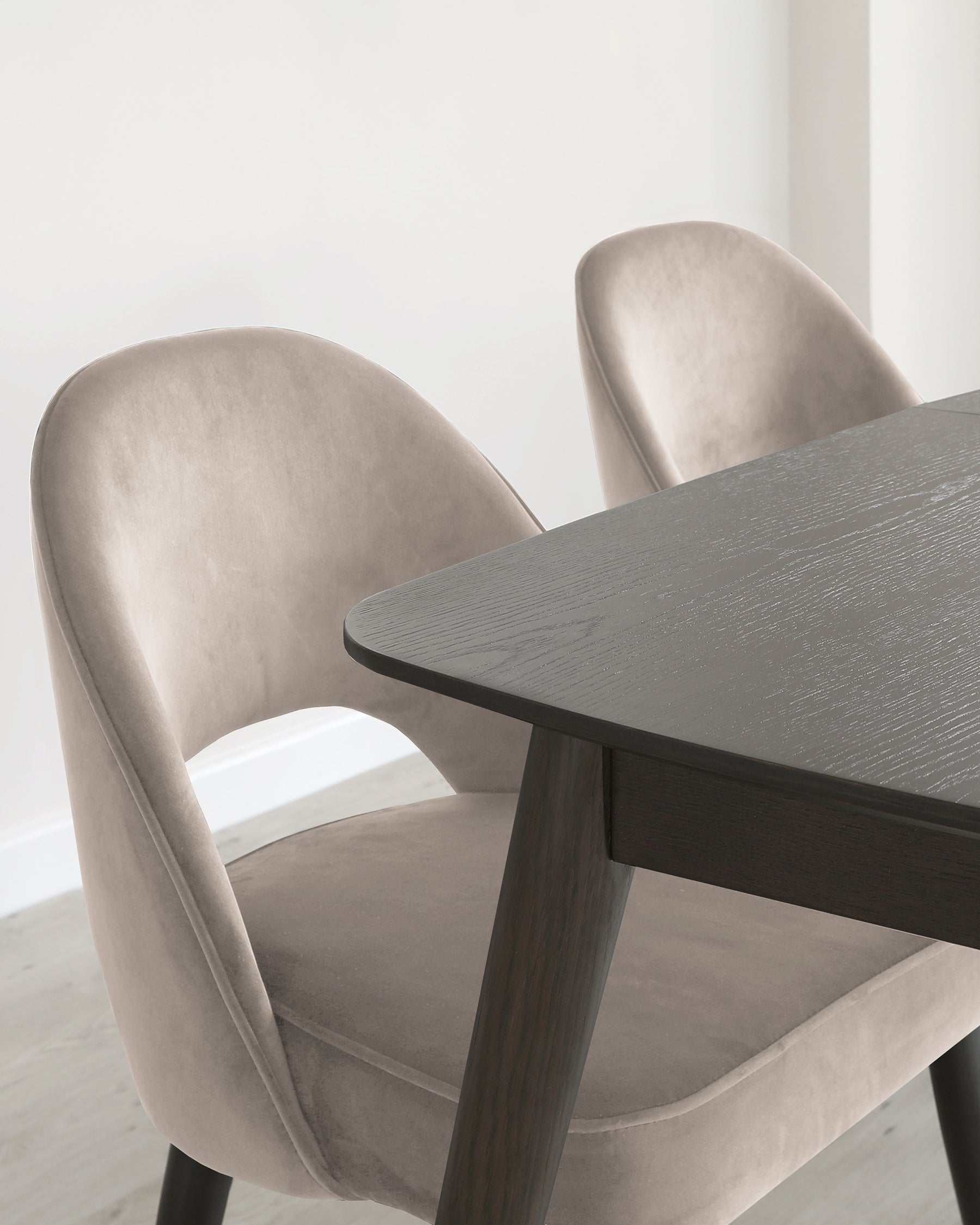 Elegant modern dining set featuring two plush, velvet-upholstered chairs in a soft taupe colour with a smooth, curved backrest and dark wooden legs. A dark wooden table with a textured surface and round, angled edges complements the chairs.