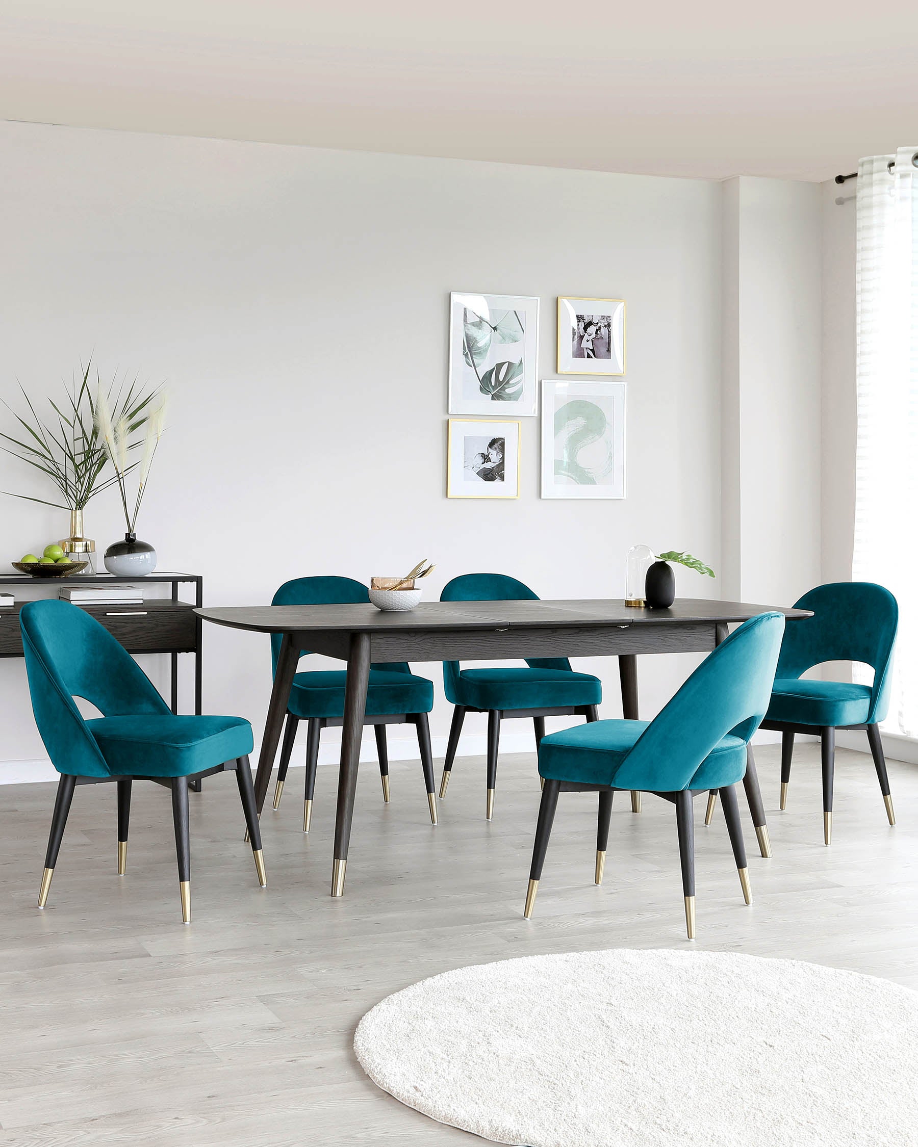 Modern dining room featuring a dark wood table with black legs tipped in gold, surrounded by six teal velvet upholstered chairs with black and gold legs. A dark wood console table is accented with decorative vases and accompanied by a circular white rug on a light wooden floor.