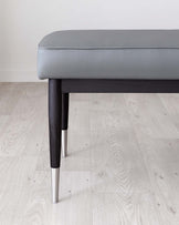 Modern minimalist bench with a sleek grey upholstered cushion and dark-stained wooden frame, featuring distinctive metal-tipped tapered legs.