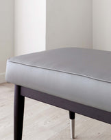 A modern minimalist style ottoman with a grey upholstered top and dark wooden frame featuring tapered legs with metallic tips.