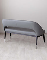 Elegant modern grey upholstered bench with a curved backrest and black legs that feature metallic tips, set against a light wooden floor and a neutral wall background.