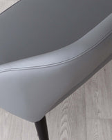 Close-up view of a modern chair featuring a smooth grey faux-leather seat with subtle stitching details. The chair has a sleek profile with a gently curving backrest and is supported by a slim, matte black metal leg, partially visible. The flooring is a light wood laminate, providing a neutral backdrop for the chair.