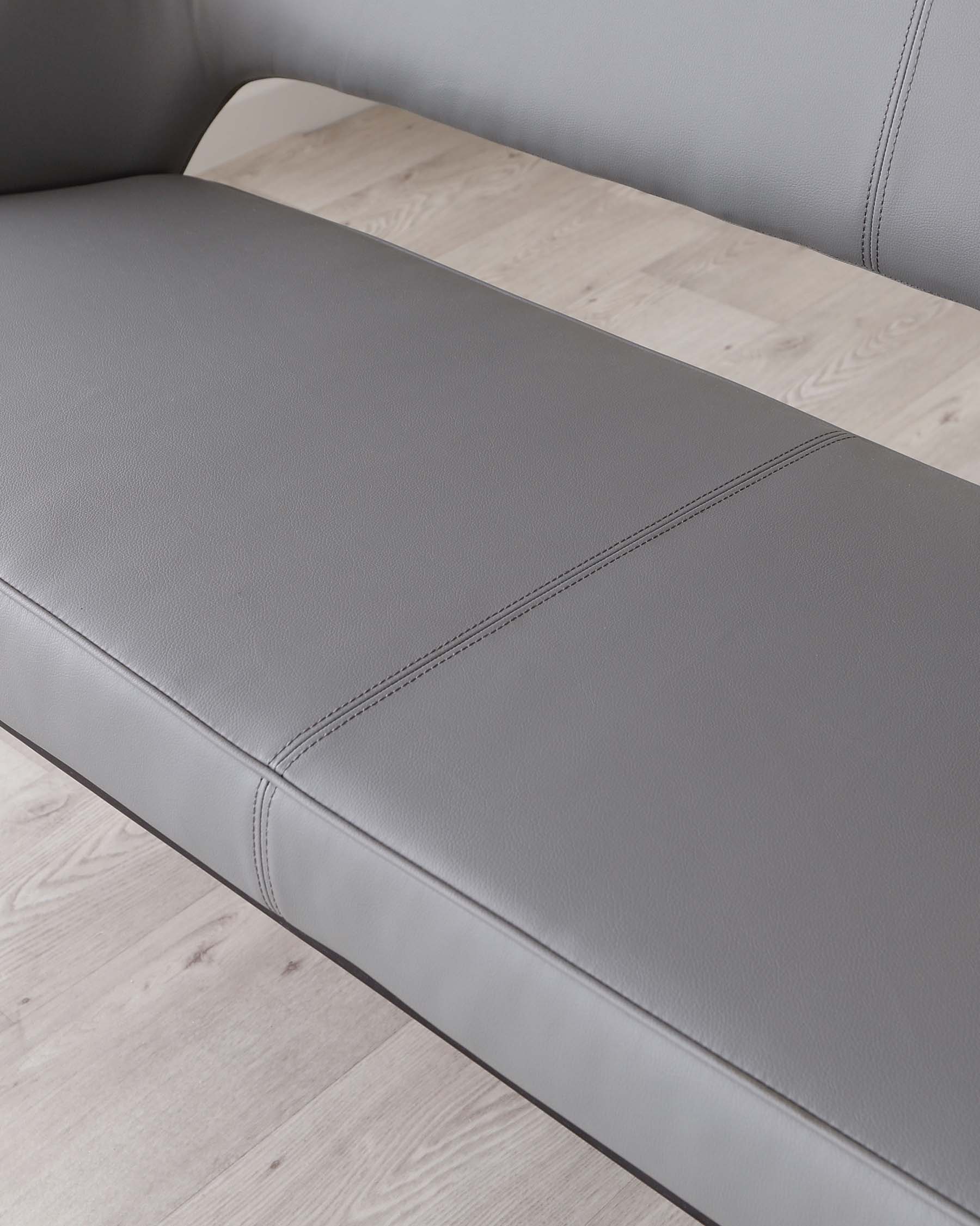 Modern sleek grey leather sofa with clean lines and minimalistic design, placed on a light wooden floor. The sofa features subtle stitching details that accentuate its rectangular form and plush seating surface.