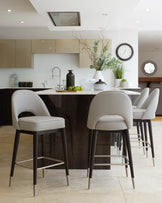 Three sleek, modern bar stools with a curved backrest and cushioned seat in light grey upholstery, featuring dark wooden legs with metallic footrests, situated at a kitchen island.