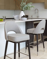 Two modern bar stools with grey upholstery and dark wooden legs, featuring a sleek design with low backrests and footrests, placed by a kitchen island.