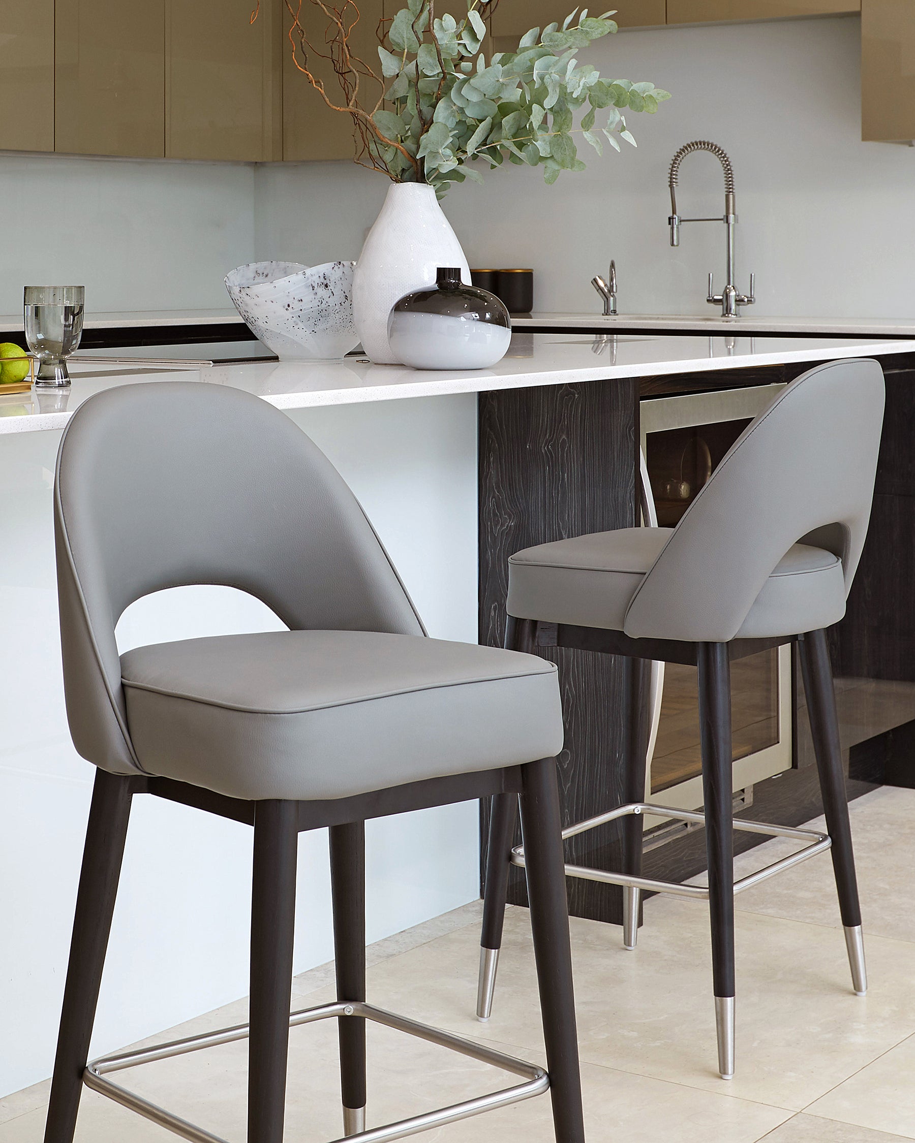 Modern bar stools with grey upholstered seats and backs, black wooden legs, and metallic footrests positioned at a white kitchen counter.