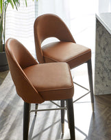 A pair of modern caramel-coloured leather upholstered dining chairs with curved backs and cushioned seats, featuring thin black metal legs, positioned near a kitchen island with a white marble countertop. The setting is bright with natural light filtering through sheer curtains.