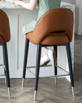 Modern barstool with a curved, camel-coloured faux leather seat and black metal legs featuring silver accents at the feet and a round footrest.