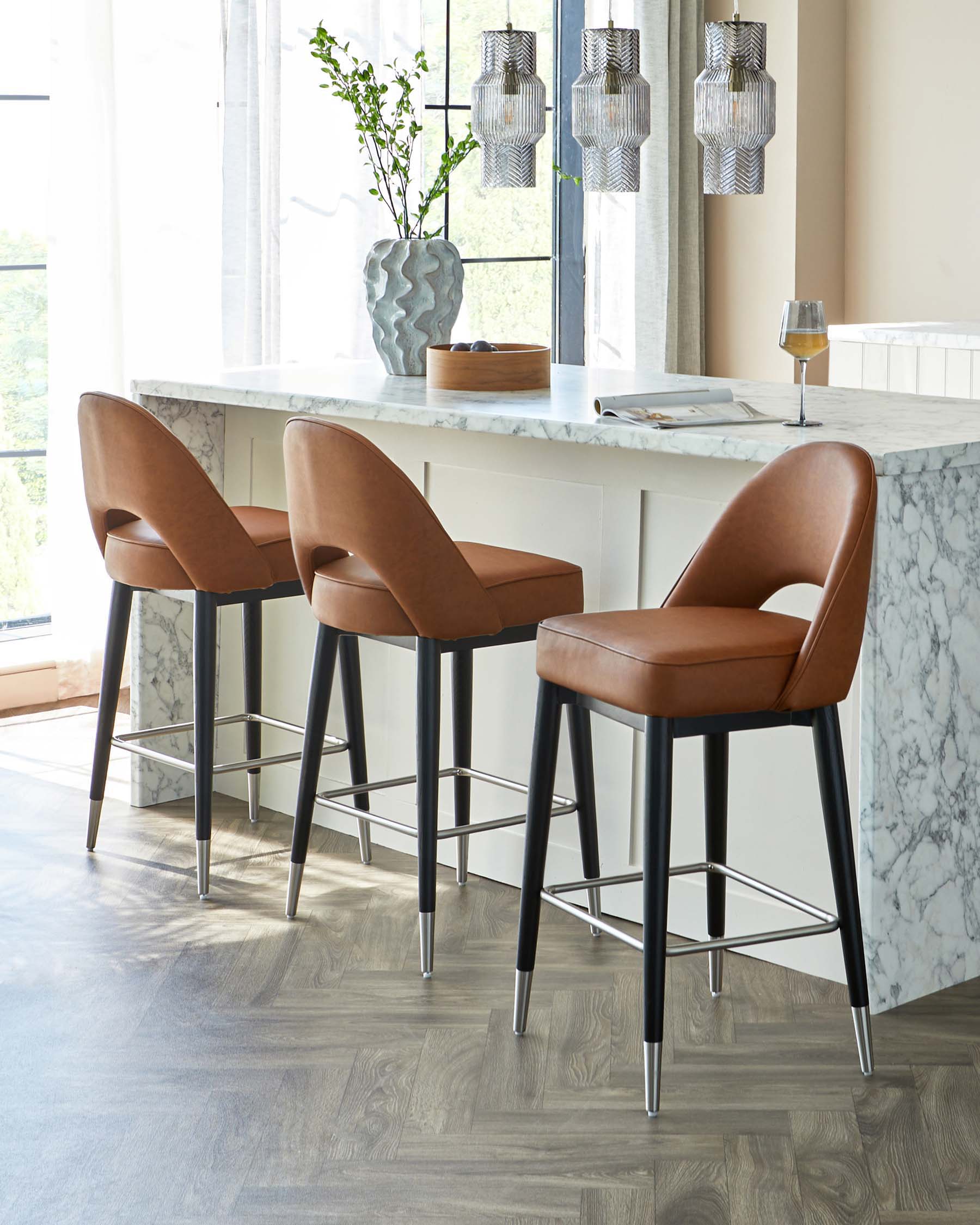 Three modern bar stools with sleek caramel brown leather upholstery, curved backrests, and slender black metal legs with built-in footrests, positioned by a white marble kitchen island.