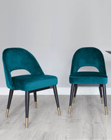 Two elegant teal velvet accent chairs with black tapered legs accented with gold tips, set against a light grey wood floor and white wall.