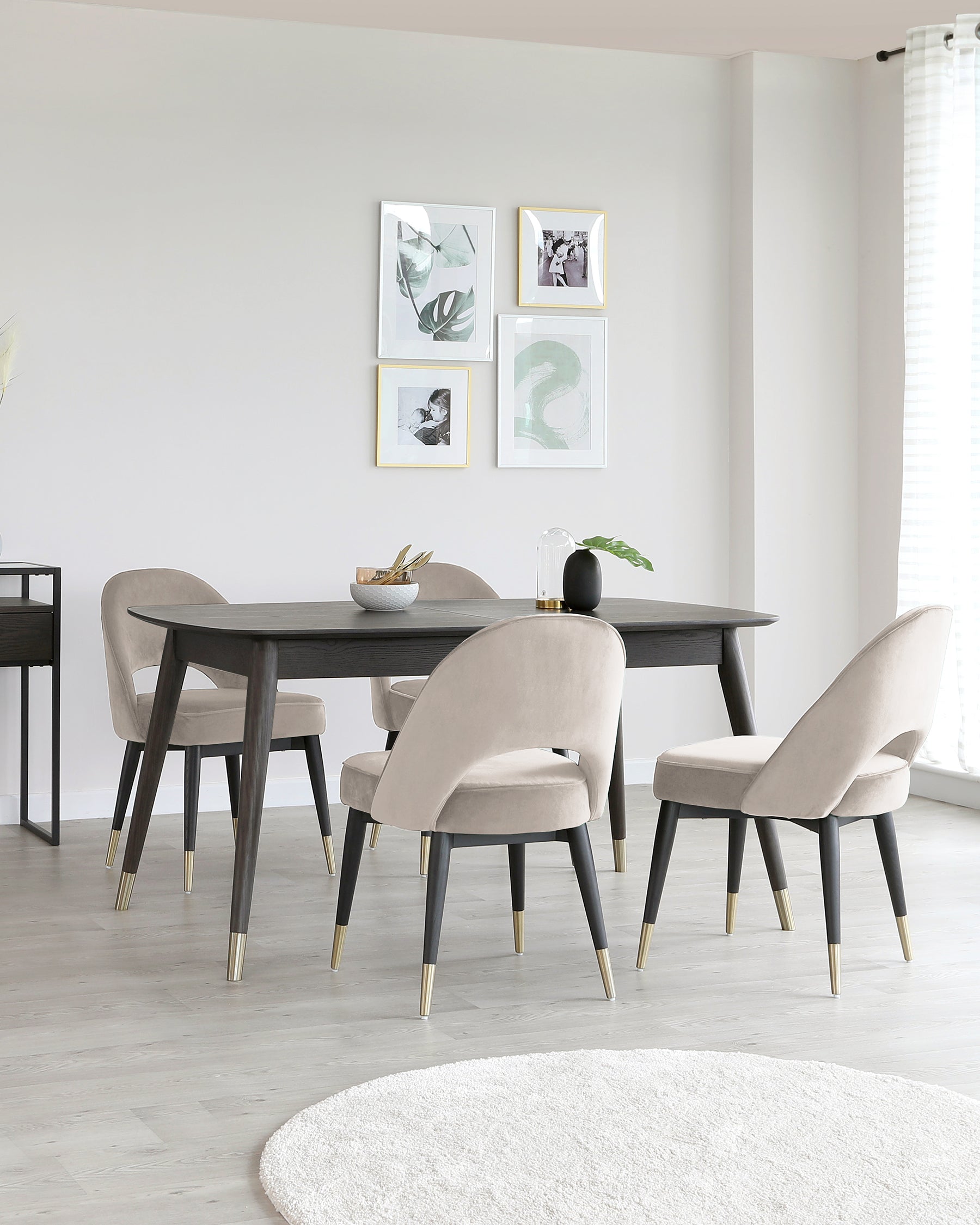 Modern dining room setup featuring an elegant rectangular dark wood table with slender legs capped by brass feet, accompanied by four plush velvet-upholstered chairs with curved backs and brass-tipped legs, on a light hardwood floor.