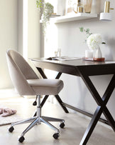 A modern home office setup featuring a black wooden desk with X-shaped legs and a beige upholstered office chair on a chrome star base with casters.