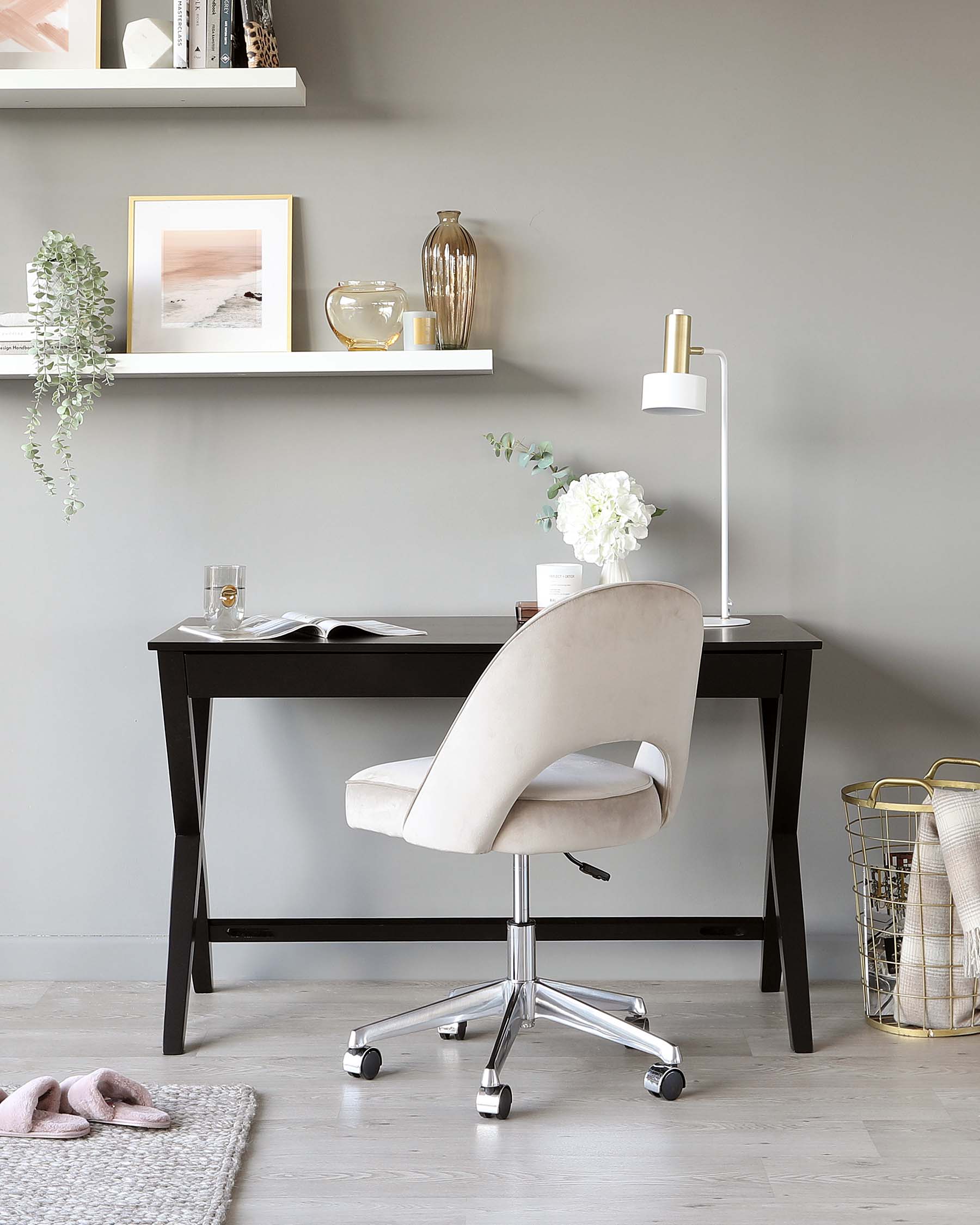 A modern home office setup featuring a sleek, black wooden desk with angled legs, paired with a contemporary light beige upholstered swivel office chair on a chrome star base with casters. The chair has a curved backrest and padded armrests for comfort. The simple and elegant design of the furniture is complemented by a minimalistic decor.