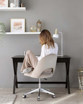 A modern, dark wooden desk with clean lines and tapered legs paired with a light beige upholstered office chair on a five-point chrome rolling base. The desk is accompanied by a thin white floating shelf above, displaying decorative items. The setup is in a minimalist grey-walled room with a light wooden floor.