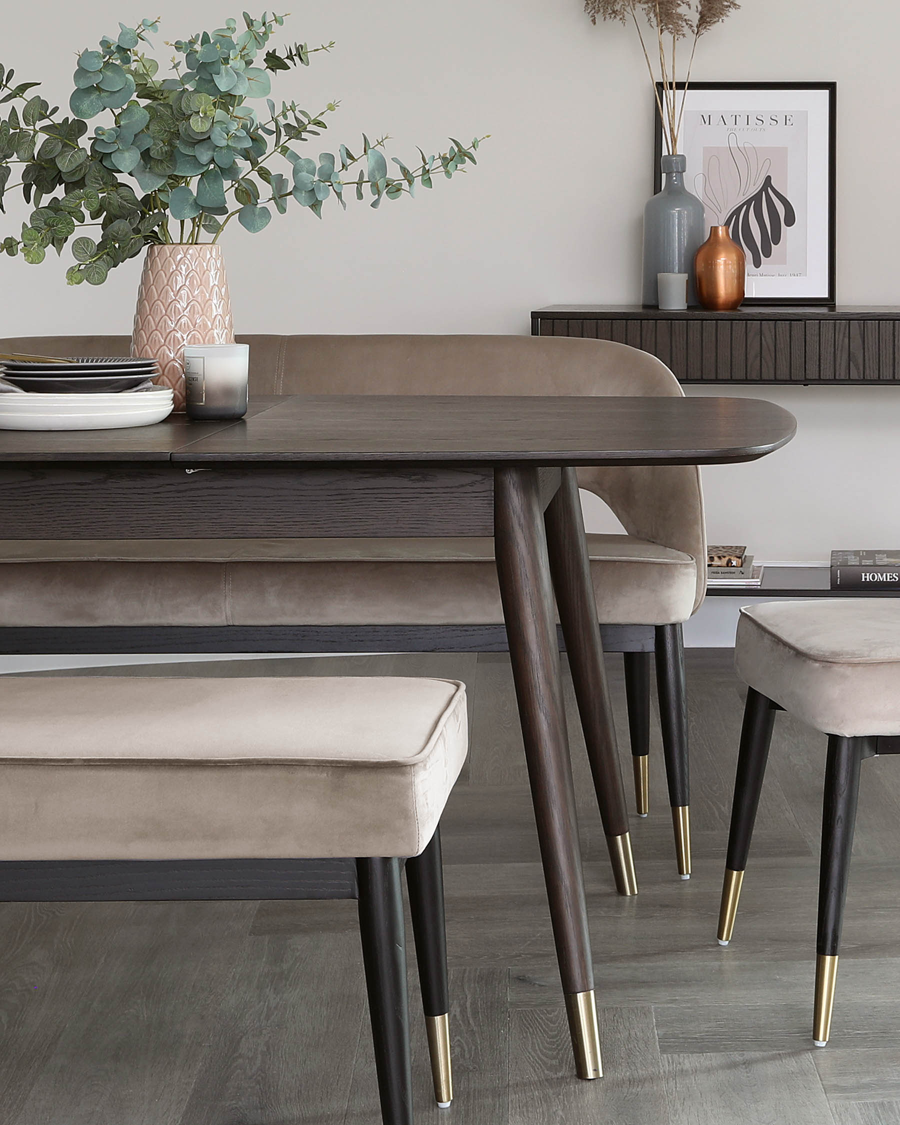 Modern dark wooden dining table with tapered legs and brushed gold feet, accompanied by two matching upholstered bench seats in a light taupe fabric, also featuring tapered legs with brushed gold feet.