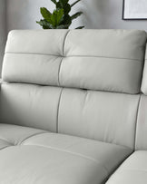 Close-up of a modern light grey leather sofa with a tufted backrest and plush cushioning. A decorative plant is subtly visible in the background, adding a touch of greenery.