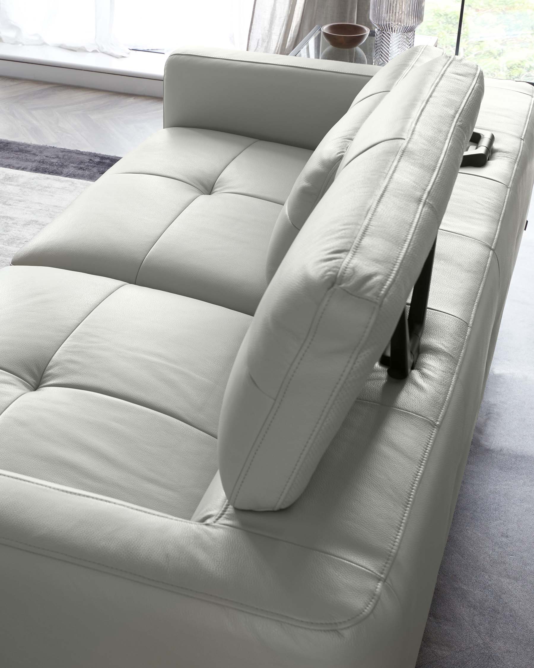 Modern light grey leather sofa with clean lines and a plush, tufted seat design, featuring adjustable headrests and a streamlined armrest with a simple lever. The couch is placed in a bright room with a floor-to-ceiling window, and hints of a translucent curtain and a fuzzy grey rug are visible, emphasizing contemporary comfort and style.