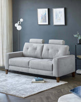 A modern light grey fabric three-seater sofa with straight clean lines, featuring cushioned backrest and seat, flanked by square armrests, raised on tapered wooden legs. The sofa is positioned on a textured off-white area rug over a hardwood floor, with a sleek black round side table to the right and an elegant white globe floor lamp to the left. Two framed abstract artworks hang on the grey wall behind the sofa, adding a touch of colour to the sophisticated, minimalist setting.