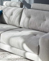 Contemporary light grey fabric sofa with textured upholstery and plush cushioning. Features a streamlined design with rounded armrests, distinctive seam detailing, and positioned on a pale floor against a high-contrast shadowed background.