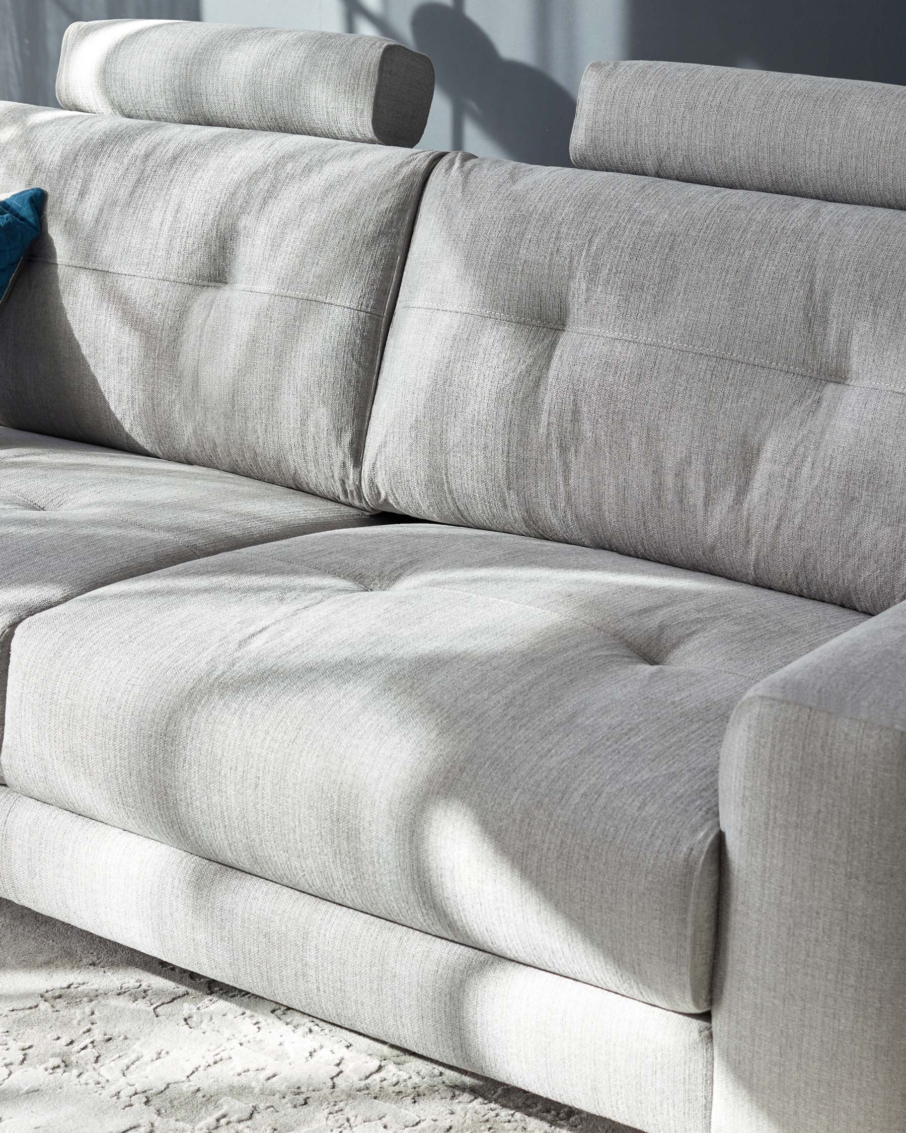 Close-up view of a section of a contemporary grey fabric sofa with a soft texture, showcasing the cushioned armrest, backrest, and seat. The sofa is illuminated by natural light, emphasizing its clean lines and comfortable design. A subtle shadow pattern adds depth and interest, while a textured area rug beneath suggests a cosy, stylish living space.