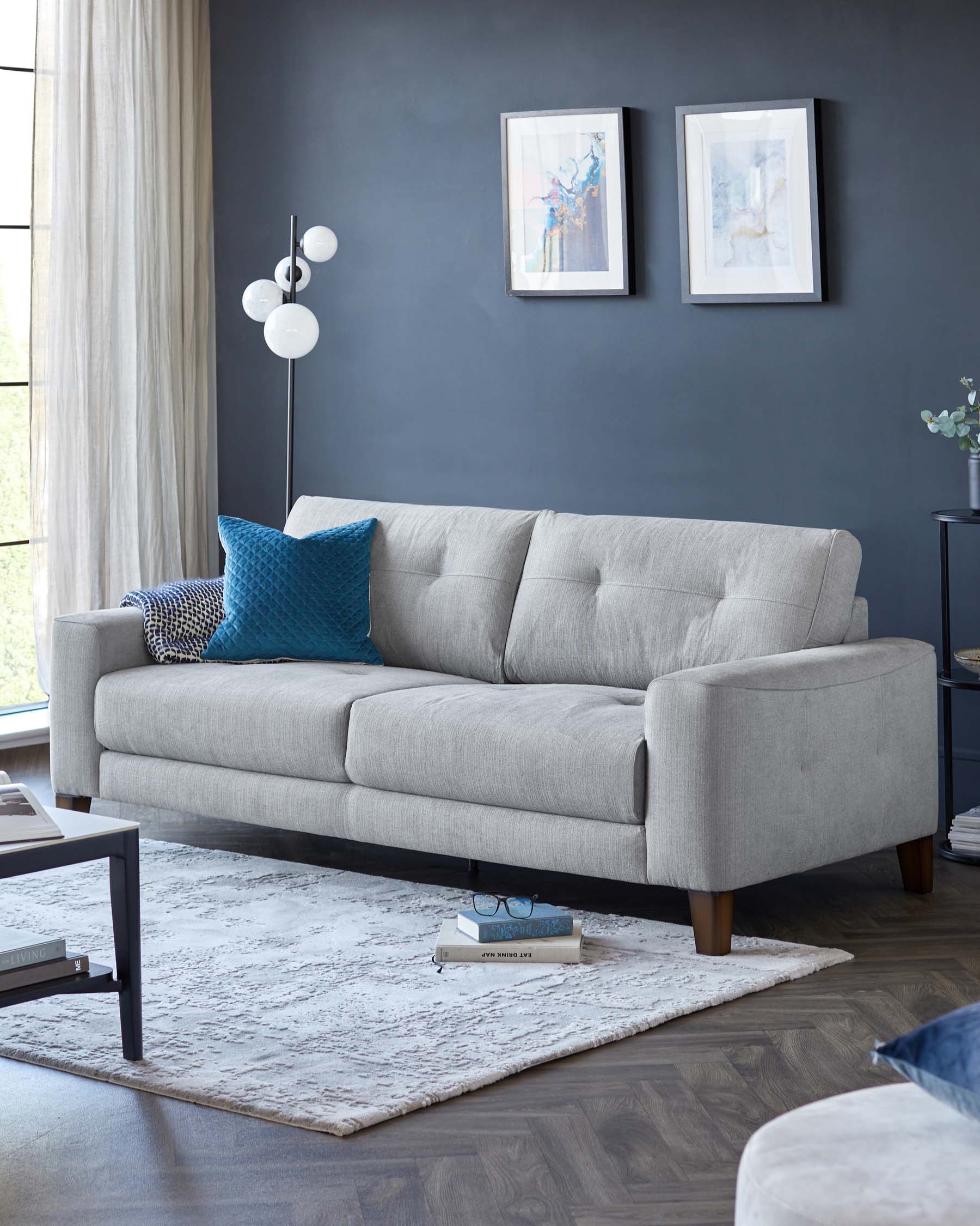 A modern light grey fabric three-seater sofa with simple lines and wooden legs, accented with various textured pillows in shades of blue and grey. In front of the sofa lies a large off-white textured area rug, and to the side stands a sleek, black end table with minimalist design.