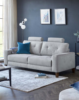 A contemporary light grey fabric three-seater sofa with a tufted back, clean lines, cylindrical wooden legs, and adorned with blue and patterned throw pillows. A chic side table with a round, dark surface next to the couch, over a textured off-white area rug. A floor lamp with white spherical shades extends behind the sofa.