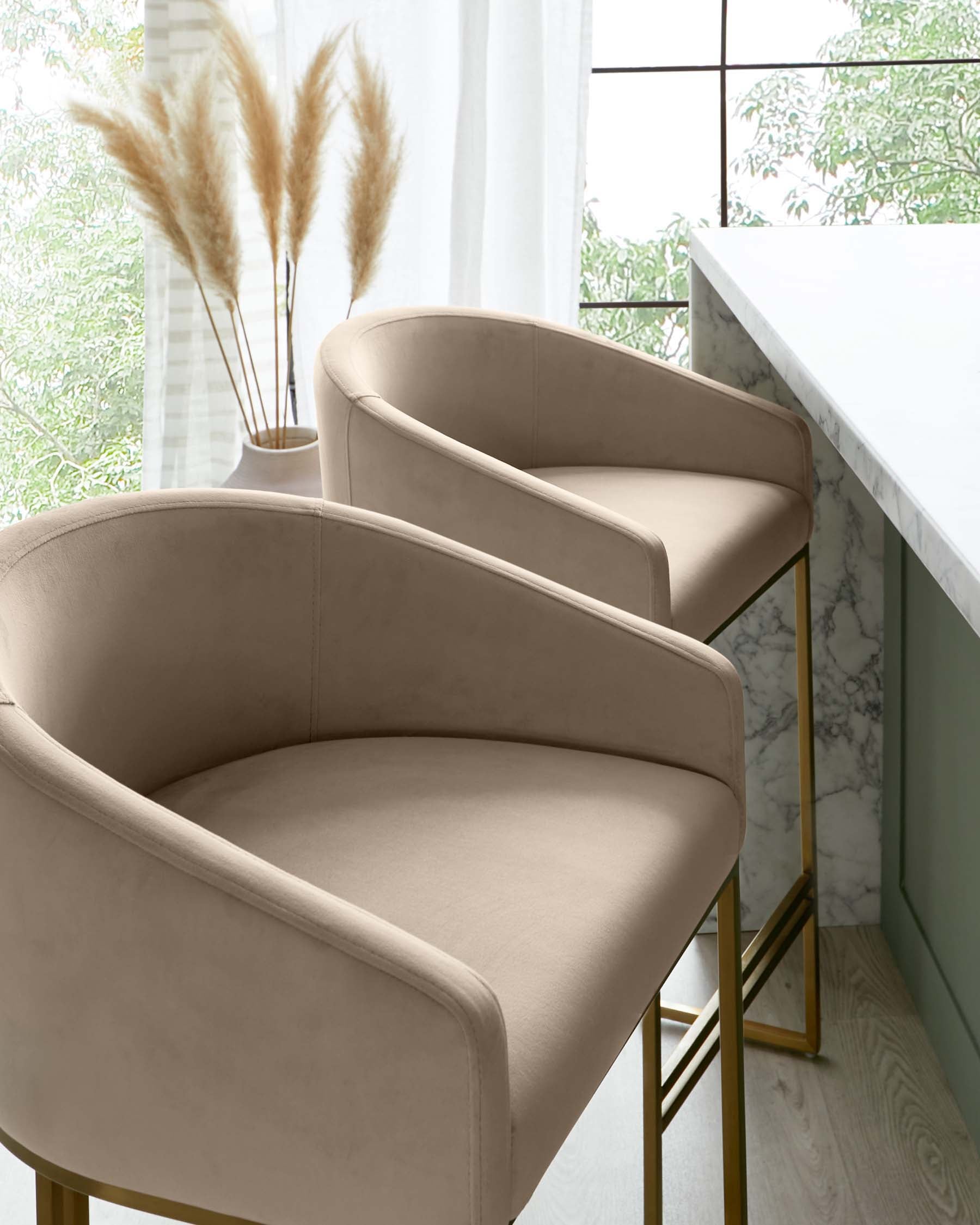 Two elegant beige upholstered bar stools with a curved back design and slim golden metal legs, positioned beside a white marble countertop.