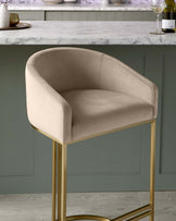 Elegant modern bar stool with a plush, beige velvet upholstered seat and a curved backrest, supported by a sleek golden metal frame with clean lines and a minimalist circular base design. The stool is positioned next to a kitchen counter with a white marble countertop.