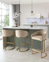 Elegant contemporary kitchen featuring a marble-topped island with a green base, adorned with three plush velvet bar stools in a soft beige colour, each with a curved backrest and sleek gold metal legs that add a touch of luxury. The island serves both as a prep area and a casual dining space, seamlessly integrating functionality with sophisticated design.