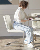 Modern ivory upholstered dining chair with a sleek chrome cantilever base, paired with a matching ivory leather upholstered bench in a well-lit room with herringbone wood flooring.