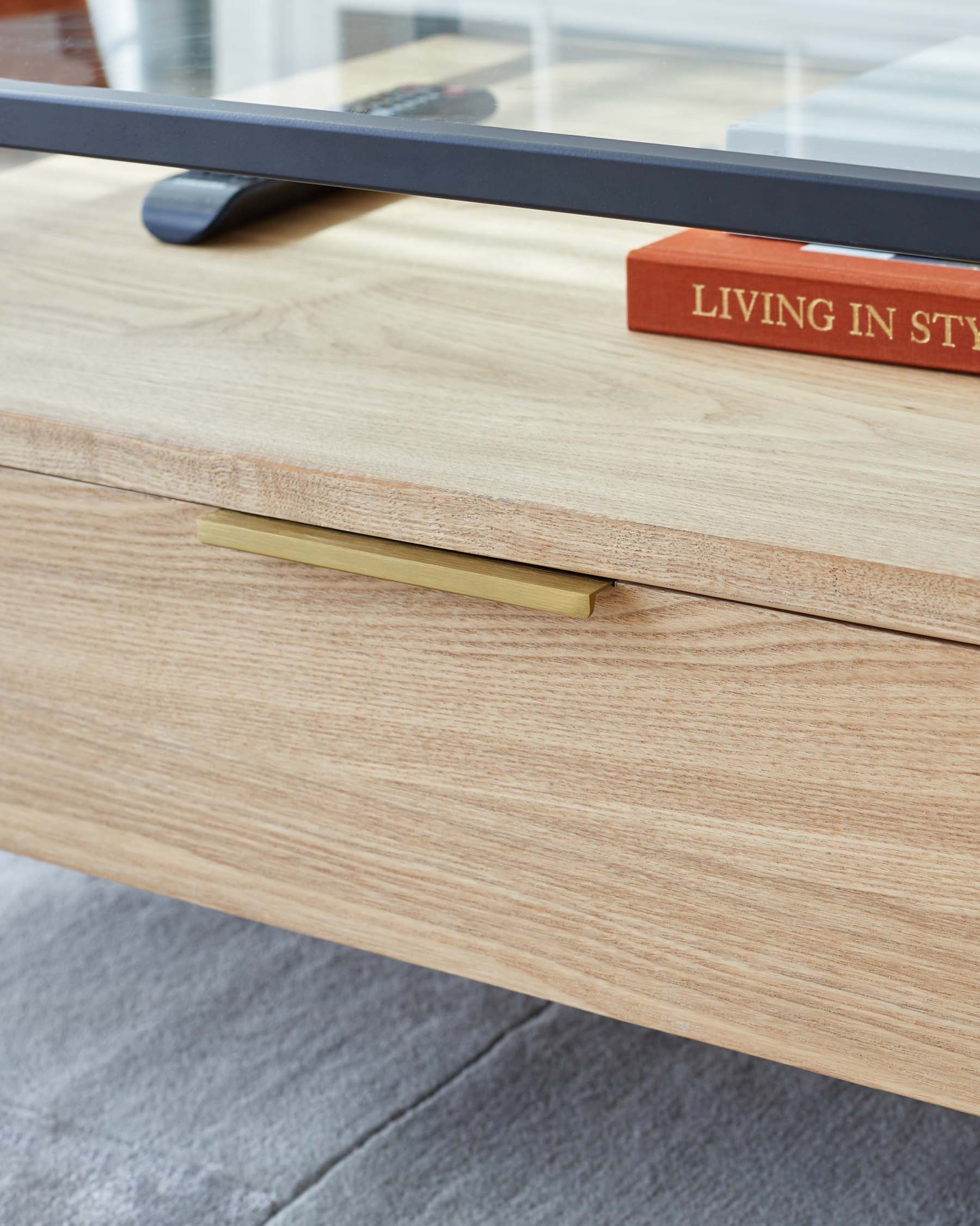 Close-up of a modern light wood coffee table with a sleek brass handle on a drawer, featuring a clear glass top through which a remote control is visible, and accompanied by a red book with "LIVING IN STYLE" on the spine, placed on the glass top. The table displays fine wood grain and is situated on a grey floor, suggesting a contemporary and clean design aesthetic.