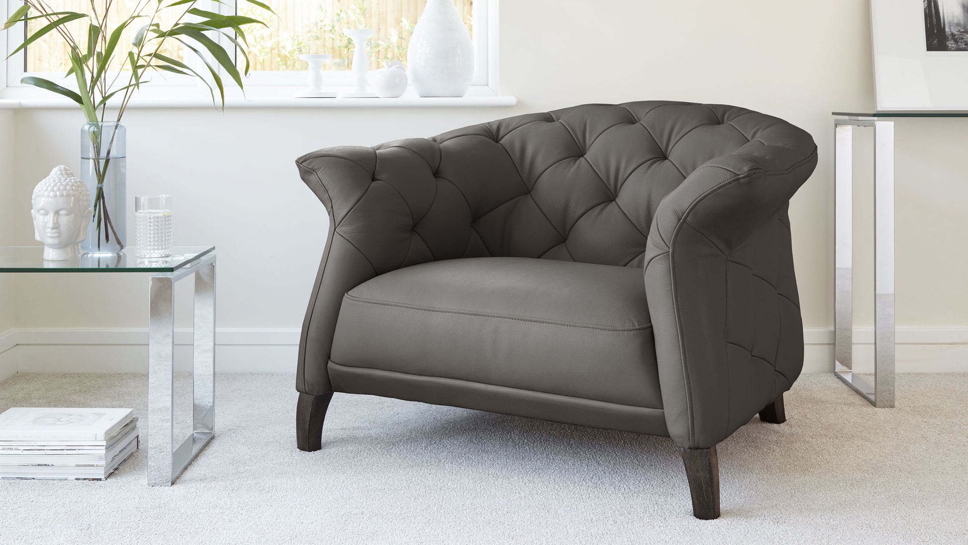 Elegant grey tufted armchair with dark wooden legs, paired with a modern clear glass side table featuring chrome legs, set against a light, neutral interior.