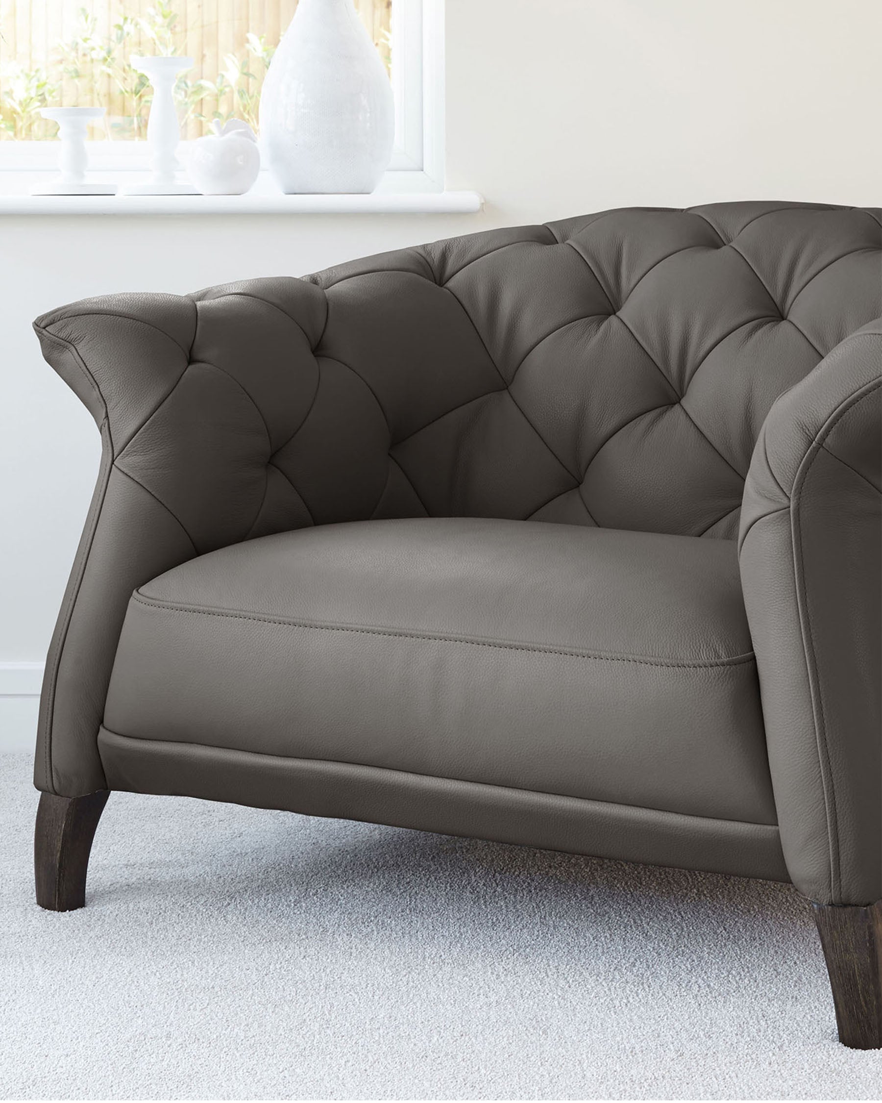 Elegant grey tufted loveseat with dark wooden legs and curved armrests, showcasing a plush back and a smooth seat, positioned on a white carpet against a light-filled room with a clear view of a window and decorative vases on the windowsill.