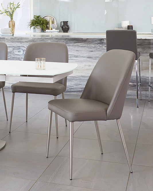 Modern dining room with a white rectangular table accompanied by sleek taupe upholstered chairs with slender metal legs. In the background, a complementary white sideboard is adorned with decorative vases and plant arrangements.