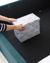 Modern geometric patterned ottoman in black and white being placed on a dark solid-colour sofa with a contrasting teal piping detail.