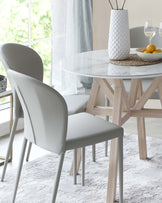 A modern dining set featuring a round marble-top table with a unique, angular wooden base, paired with sleek, grey upholstered chairs with light wooden legs. The scene is completed with a textured white vase on the table and soft, natural light filtering through sheer curtains.