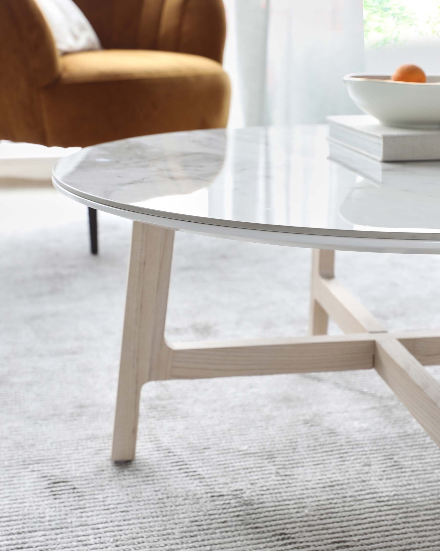 A modern round coffee table with a white marble top and Scandinavian-style pale wooden legs, placed on a textured light grey rug. In the background, there's a partial view of a mustard yellow velvet armchair with a round cushion and dark tapered legs.