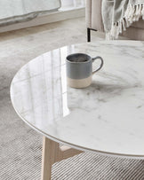 Round marble-top coffee table with a natural wood base placed on a textured off-white rug, accompanied by a two-tone ceramic mug on top.